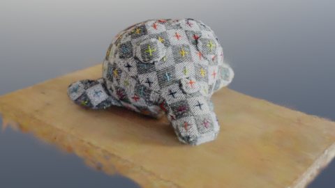 A rendered Gaussian-splat scan of a stuffed Suzanne monkey head, covered in grey patchwork fabric with coloured cross-stitches, sitting on a wooden board against a grey-blue backdrop.
